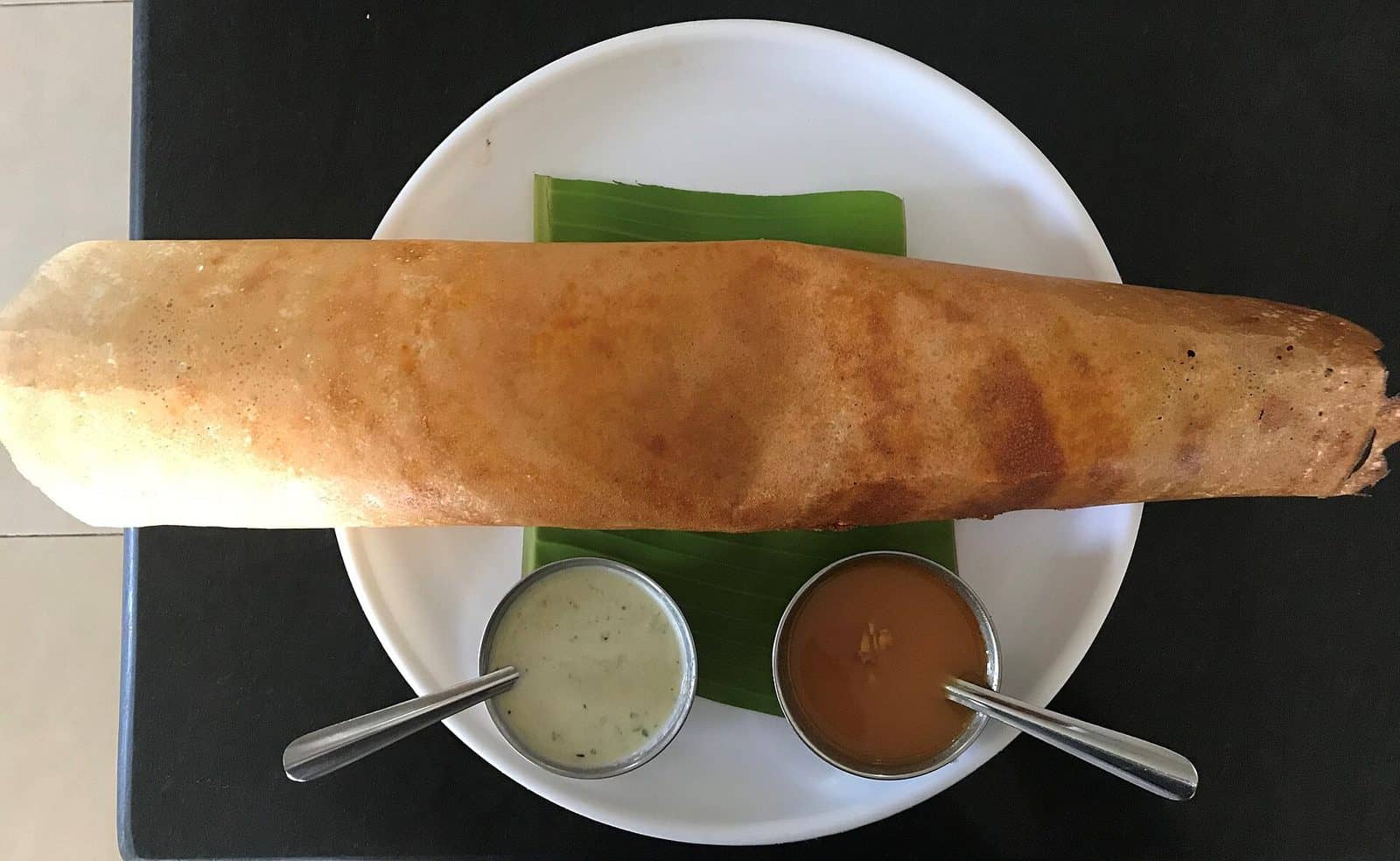 A golden rolled masala dosa on a banana leaf with coconut chutney and sambar